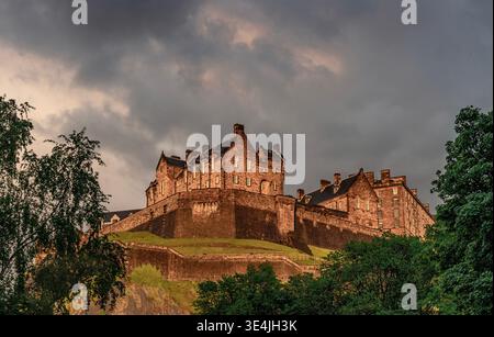 Historic Edinburgh Castle sits on a rocky crag, warmed by late evening light and framed by dramatic clouds and leafy trees, highlighting centuries-old Stock Photo