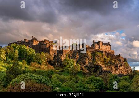 Dramatic view of Edinburgh Castle perched on Castle Rock at golden hour, bathed in warm light against stormy clouds with lush green trees and rugged c Stock Photo