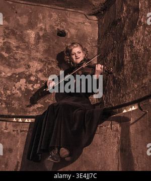 A female fiddler in vintage costume plays violin by candlelight in a weathered West Bow passageway, delivering a dramatic, moody street theatre festiv Stock Photo