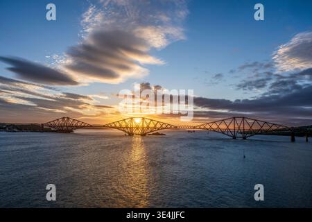 Sunrise illuminates the iconic Forth Rail Bridge across the Firth of Forth, casting golden reflections on the water beneath dramatic morning clouds an Stock Photo