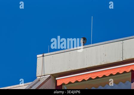 A security camera installed on the rooftop of a building under a bright blue sky, featuring a red awning and concrete structure. Stock Photo