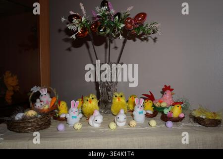 Easter table decoration with chicks, hens, rabbits and colorful eggs arranged around a glass vase with flowers, festive spring home display. Stock Photo