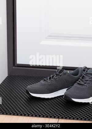 A pair of dark grey athletic sneakers is prominently featured, resting on a robust black grid-patterned rubber doormat. These contemporary shoes exhib Stock Photo