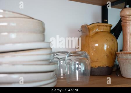 Stacked ceramic plates, glass cups and ceramic jug on kitchen shelf, neutral interior with wooden surface and kitchen storage arrangement. Kitchen Stock Photo