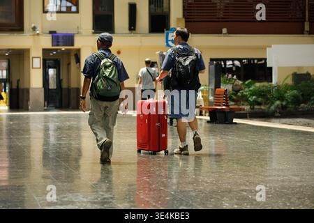 BANGKOK, THAILAND-MAR 15, 2026: Male backpackers walking and dragging luggage at a train station at Hua Lamphong, ordinary daily activity, rear view Stock Photo