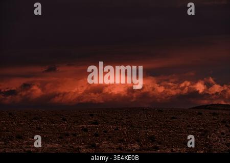 Dramatic fiery storm clouds glowing over dark desert landscape at sunset. Intense atmospheric scene with red sky, powerful weather and moody light, id Stock Photo