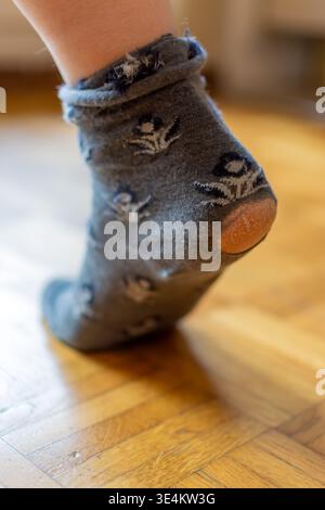 Close up of a gray sock with a hole at the heel, showing exposed skin and fabric wear against a wooden floor background. Stock Photo