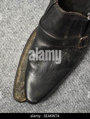 broken black leather shoe on gray carpet floor surface, worn leather material with detached front sole, top view closeup, concept of damaged footwear Stock Photo