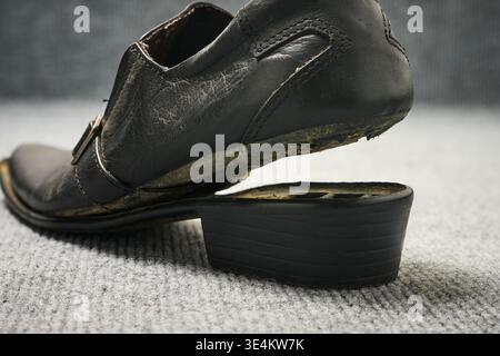 broken black leather dress shoe on gray carpet floor surface, worn leather texture with visible glue residue and detached heel, closeup side view Stock Photo