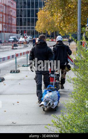 Two construction workers walk along a sidewalk at a roadwork site with barriers and an equipment cart, with autumn trees and modern buildings behind. Stock Photo
