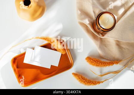 Overhead view of two blank white business cards mockup on a glossy orange ceramic tray next to a candle and dried pampas grass on a white table with a linen napkin. Stock Photo