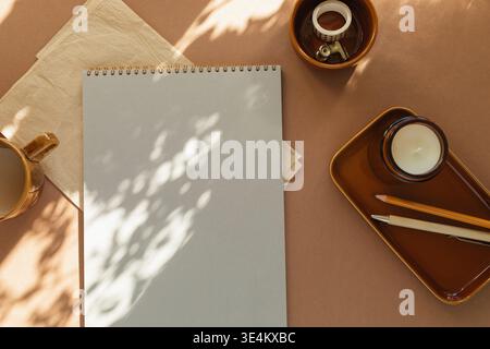 Overhead view of a blank notebook, cup of white coffee, scented block candle, pens, washi tape and a folded napkin on a table in sunlight Stock Photo