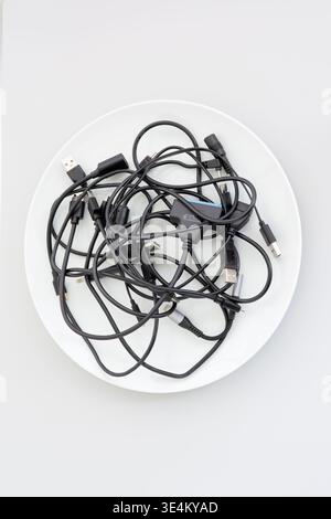 Top view of a white bowl filled with tangled black cables and assorted connectors on a light gray background. Stock Photo