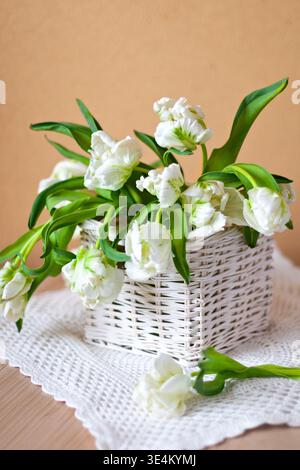 A charming arrangement of white tulips in a woven basket placed on a textured cloth, set against a warm neutral background for a cozy, minimalist stil Stock Photo