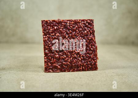 A rectangular slice of dark red fruit leather or raw seed cracker covered with visible flax seeds stands upright on a beige textured surface. The Stock Photo