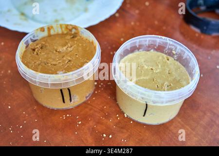 Two transparent plastic containers filled with thick nut butter sit on a reddish-brown wooden table scattered with small white crumbs. The left Stock Photo