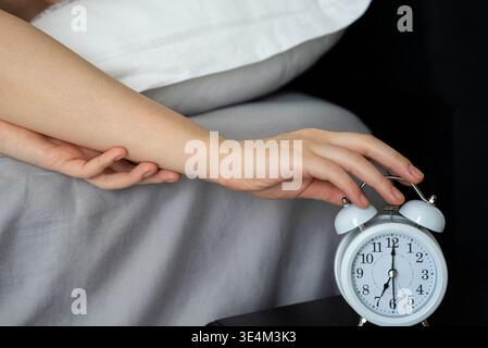 A woman in bed hits the snooze button on her alarm clock, struggling to wake up in the morning, conveying a sense of sleepiness and reluctance to start the day. Stock Photo