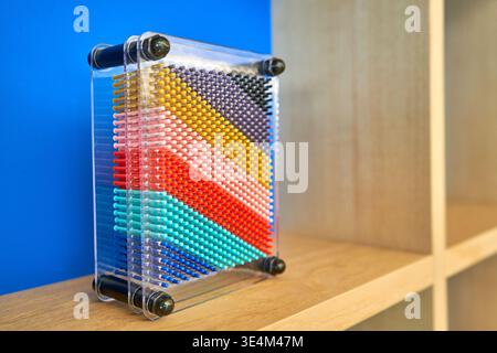 A colorful pin art impression toy displayed on a light wooden shelf. The rectangular desktop novelty features a transparent acrylic frame with black Stock Photo