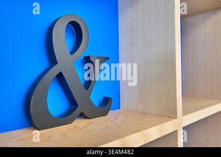 A large, dark gray decorative ampersand symbol stands on a light wooden shelf against a vibrant blue wall. To the right, a wooden bookcase with Stock Photo