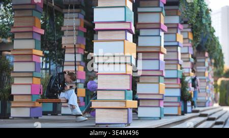 Moscow, Russia - August 17, 2025: Young woman writing on swing surrounded by colorful book columns in urban park Stock Photo