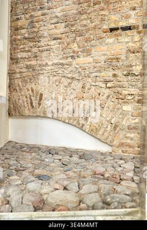 An interior view of an ancient structural brick archway and cobblestone floor. The weathered brick wall features a curved vault with bricks arranged Stock Photo