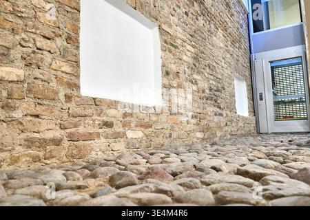 Low-angle view of a historic cobblestone pathway alongside a weathered brick wall with white-painted window frames. The foreground features smooth, Stock Photo