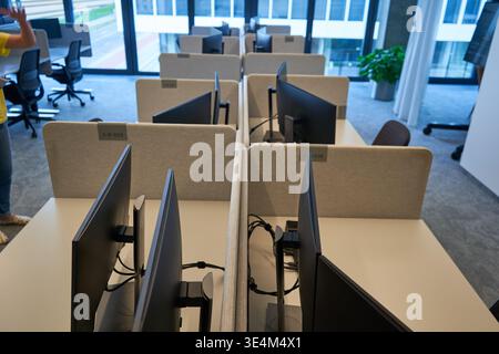 A modern office interior featuring multiple empty workstations arranged in parallel rows. Each desk is equipped with computer monitors mounted on Stock Photo
