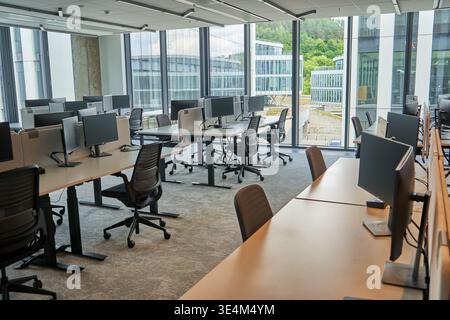 A modern, open-plan office interior featuring multiple rows of wooden desks equipped with computer monitors and ergonomic black mesh office chairs. Stock Photo