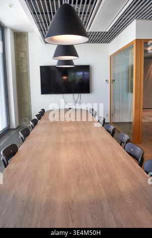 Modern conference room interior featuring a long wooden meeting table with dark gray chairs arranged on both sides. Three large black cone-shaped Stock Photo
