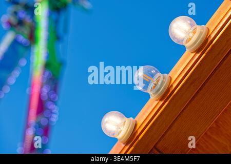 Three clear light bulbs mounted on a wooden frame against a bright blue sky with colorful blurred lights in the background creating a festive atmosphe Stock Photo