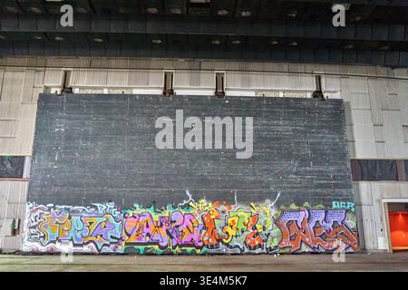 Interior view of a large abandoned industrial hall or former cinema with a massive dark projection screen or wall dominating the frame. The lower Stock Photo