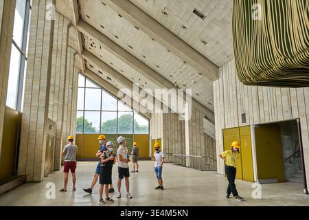 A group of people wearing safety helmets tours the interior of a large brutalist architectural space featuring exposed concrete walls with vertical Stock Photo