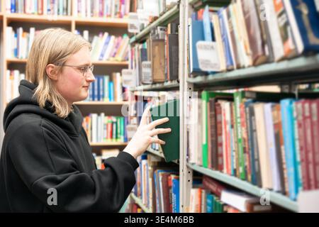 Young man browsing bookshelves in library. Smart student in glasses in library, education concept. Rows of books around. Stock Photo