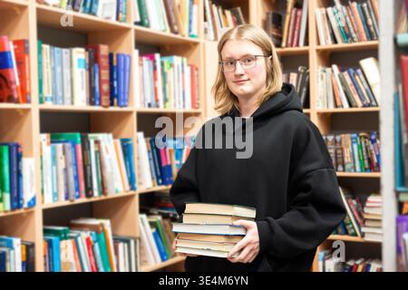 Young smiling man in a library holding stack of books. Smart student in glasses in library, education concept. Reading, learning and academic man nerd Stock Photo
