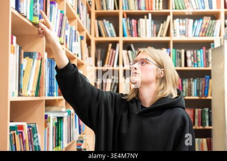 Young man browsing bookshelves in library. Smart student in glasses in library, education concept. Rows of books around. Stock Photo