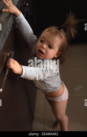 candid intimate portrait of a curious toddler in a home kitchen reaching for a cabinet handle, warm muted browns and grays, soft shadows, tiled floor, Stock Photo