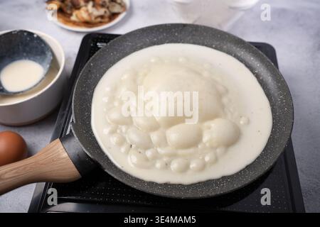 close-up of creamy white batter bubbling and puffing in a speckled frying pan on an induction cooktop, surrounded by an egg, batter bowl and blurred p Stock Photo