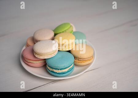 assorted pastel macarons piled on a white porcelain plate on a light wood tabletop, softly lit with shallow focus and gentle bokeh, creating an elegan Stock Photo