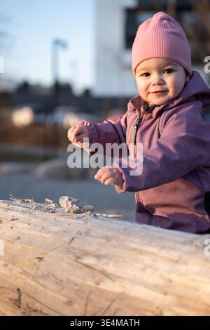 smiling toddler in a pink beanie and purple jacket playing with pebbles on a sunlit log in an urban park at golden hour candid close-up with warm tone Stock Photo
