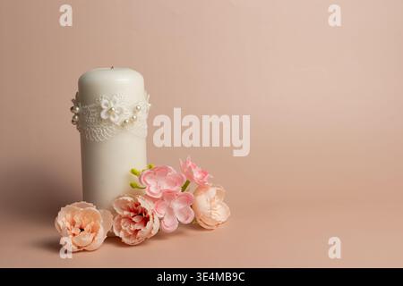 A white decorative candle with lace and pearl details is displayed with several light pink artificial flowers on a beige background with copy space on Stock Photo