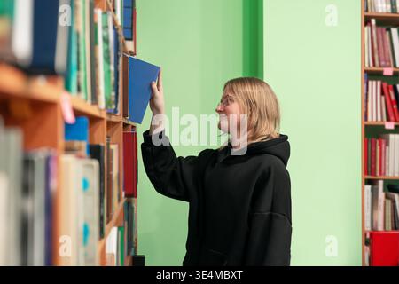 Young smiling man in a library taking book from shelf. Smart student in glasses in library, education concept. Tall shelves with books around. Stock Photo