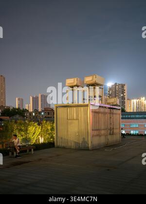 Wuhan, China - June 11, 2025: Small corrugated metal utility structure situated on a paved public plaza at night. The image shows a person sitting on Stock Photo