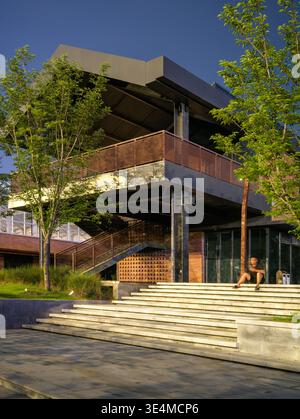 Wuhan, China - June 11, 2025: Person sitting on the wide concrete stairs of a modern architectural building at night. The image features illuminated s Stock Photo