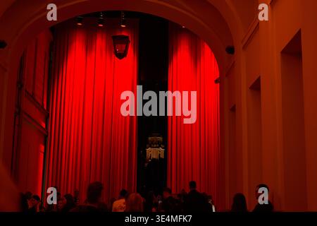 A nighttime scene showing a large crowd gathered under a grand stone archway draped with heavy red curtains that are partially drawn open, revealing Stock Photo