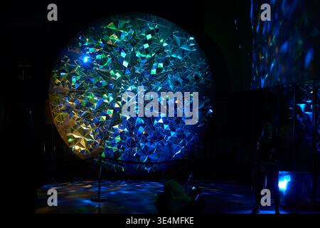 A large circular light art installation featuring a geometric crystalline structure composed of numerous triangular facets, illuminated with vibrant Stock Photo