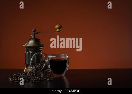 A hot coffee drink in a double-walled glass cup on a dark brown wooden table, next to a manual grinder and another cup with coffee beans spilling out. Stock Photo
