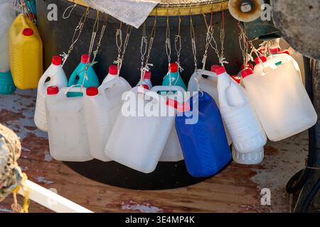 A group of white, blue, and yellow plastic jerrycans hanging from ropes on a fishing boat. Close-up of maritime equipment in a harbor at sunset. Stock Photo