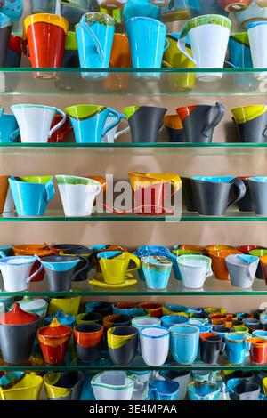Assortment of handmade colorful ceramic mugs, cups, and jugs on glass shelves. Artistic pottery with vibrant glaze in a Greek souvenir shop. Stock Photo