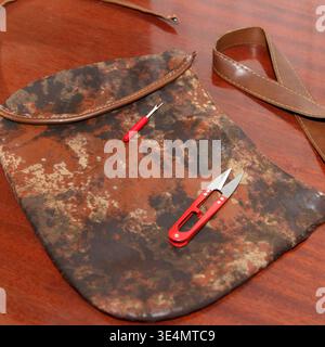 A worn, brown patterned bag is dismantled for repair on a wooden table. Red-handled thread snips and a seam ripper lay on top, ready for sewing or upc Stock Photo