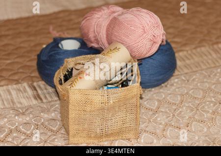A rustic still life of crafting supplies. Pink and blue balls of yarn sit behind a woven burlap basket containing a scroll. Warm, soft lighting create Stock Photo
